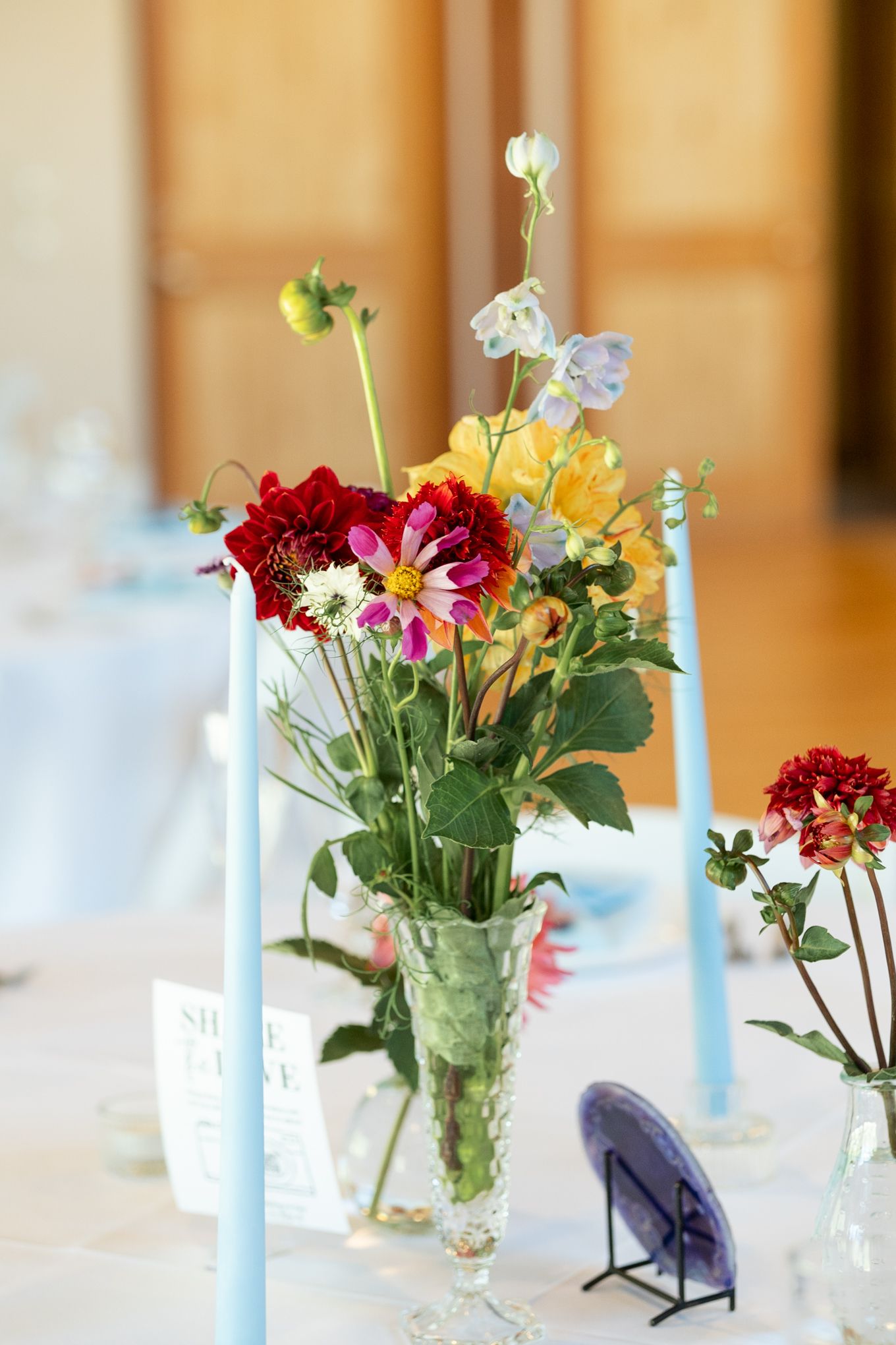 Colorful wildflower centerpiece with light blue taper candles on white tablecloth at wedding reception.