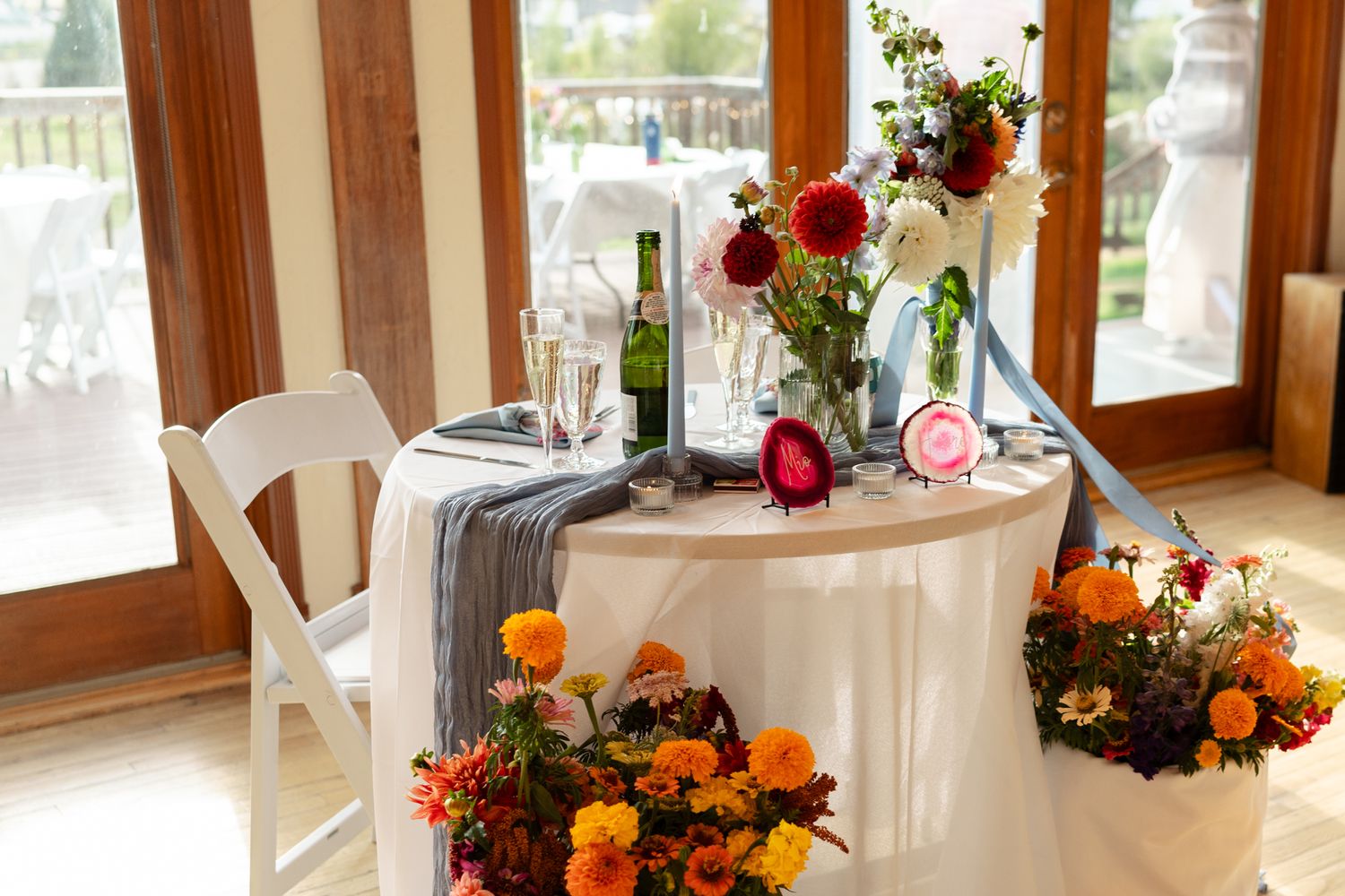 Intimate sweetheart table decorated with autumn flowers and white linens by large windows.