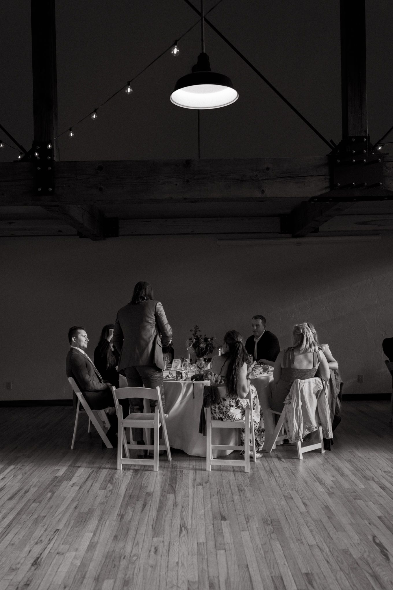 Black and white photo of wedding guests dining under industrial pendant lights in a spacious hall.