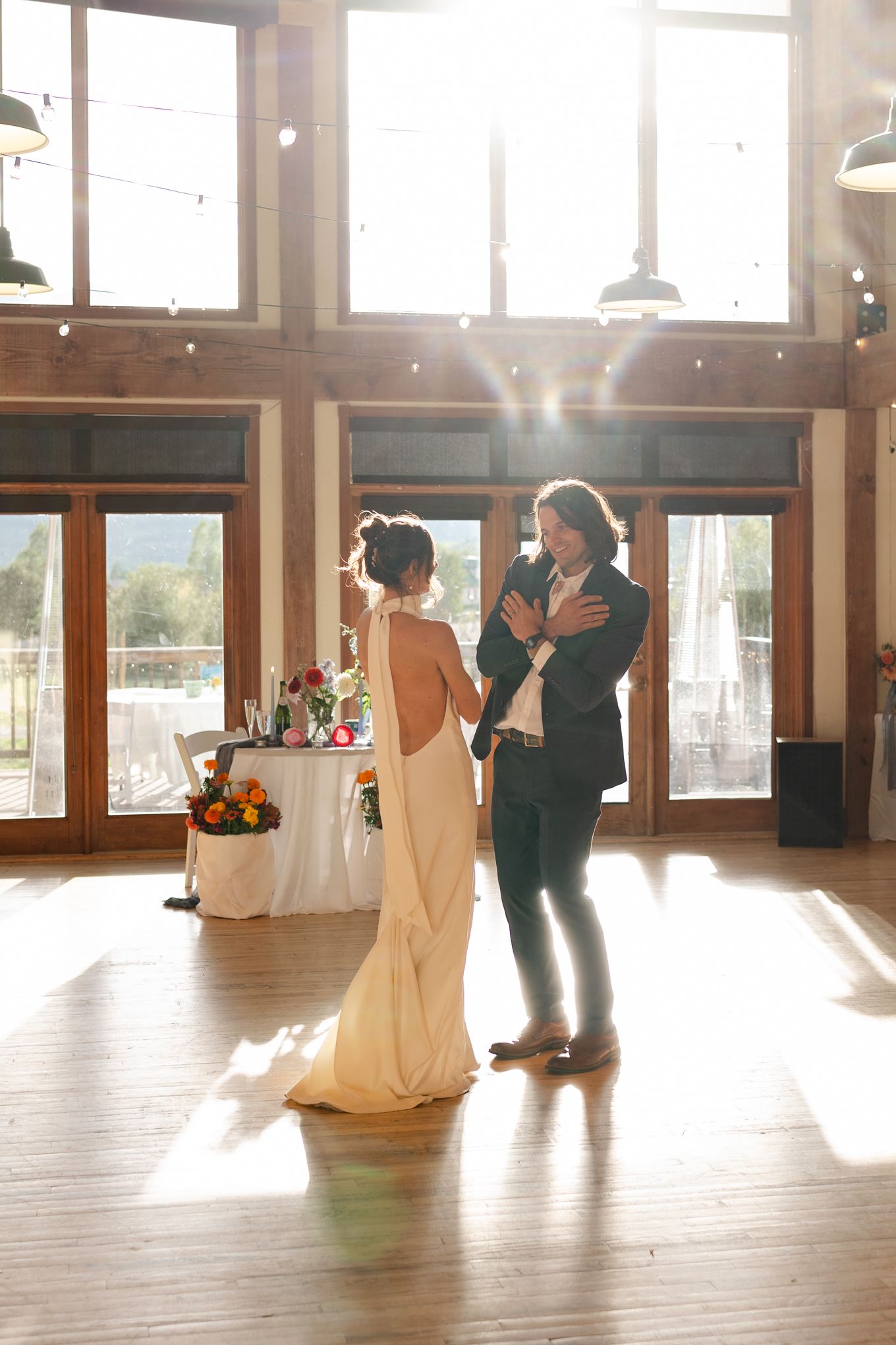 A couple shares their first dance in a sunlit venue with large windows and hardwood floors.