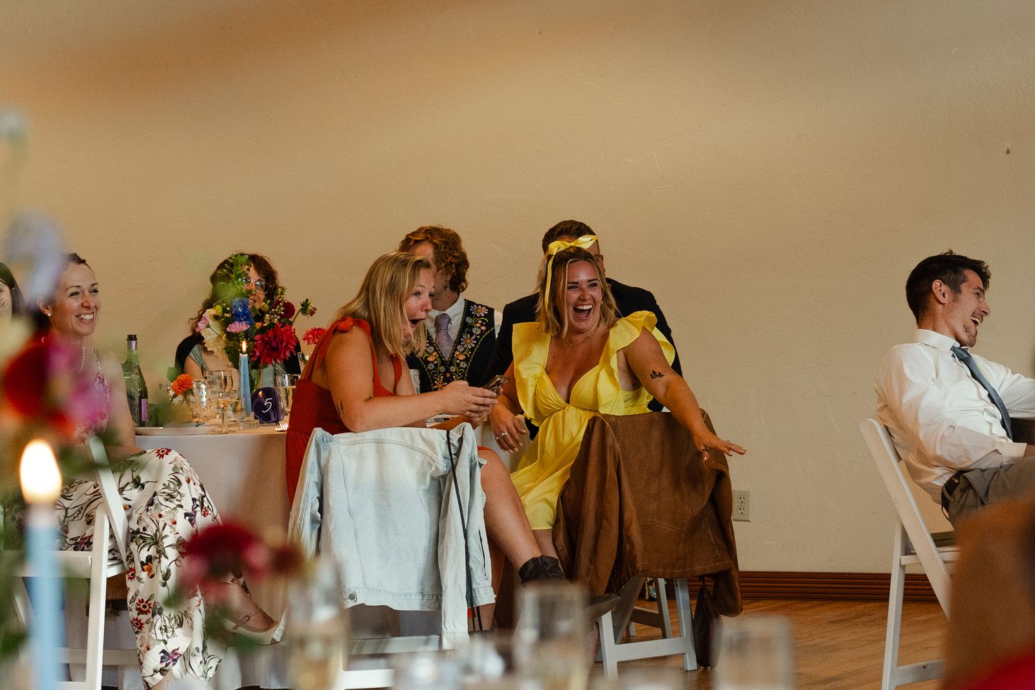Wedding guests laugh and socialize at reception tables decorated with floral centerpieces.