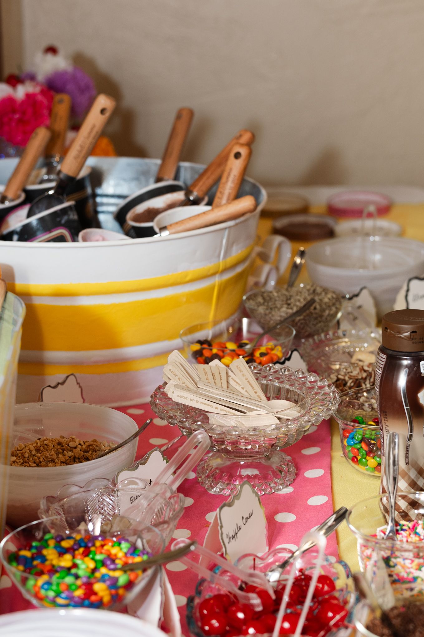 Ice cream sundae bar setup with colorful toppings and serving utensils in yellow striped containers.