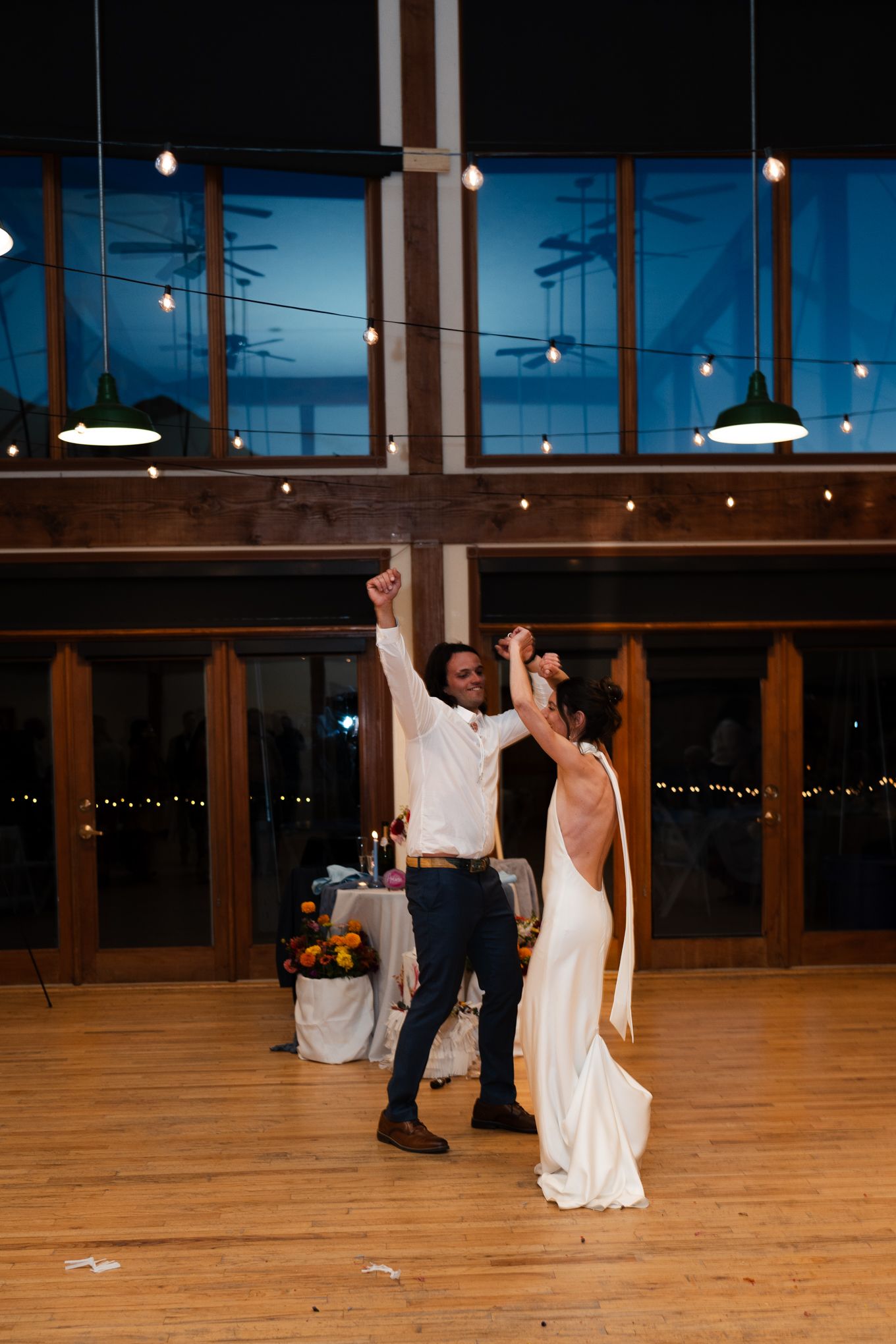 A couple dances together in an elegant venue with wooden beams and string lights overhead.