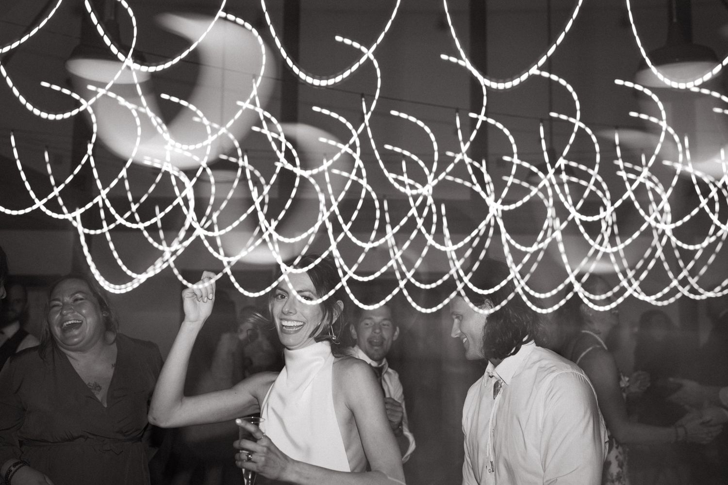 Black and white photo of party guests dancing under swirling string light decorations.