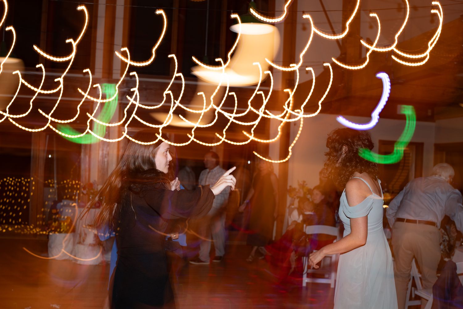Motion blurred photo of wedding reception dancers under swirling string lights in a rustic venue.