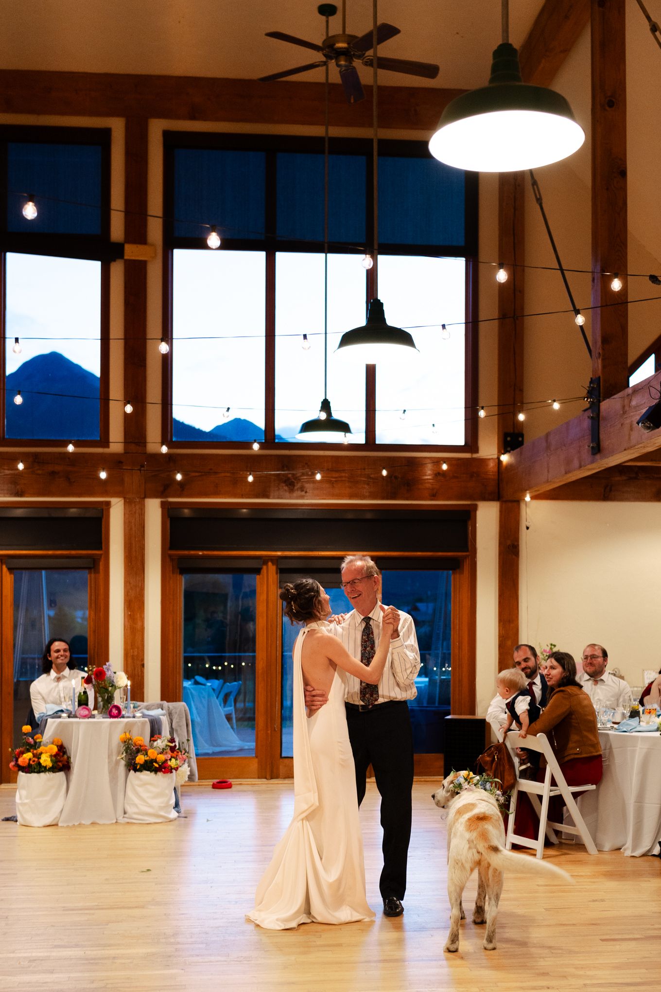 Couple dancing in an elegant venue with large windows showing mountain views at dusk while their dog watches.