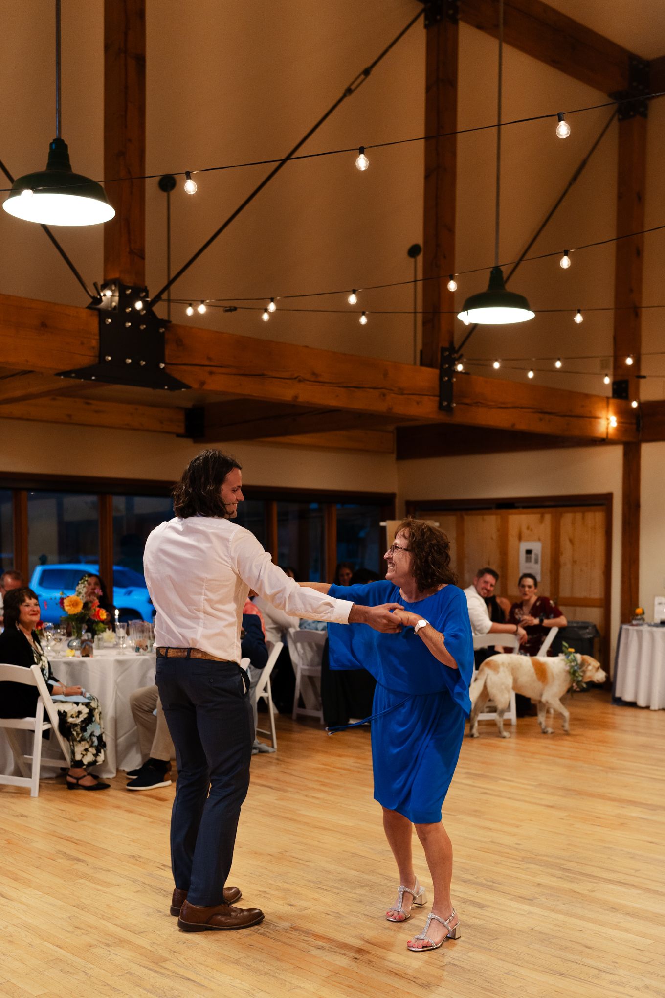 A mother and son dancing together in a rustic reception hall with string lights and wooden beams overhead.
