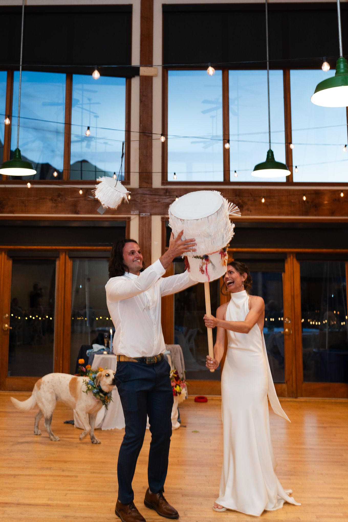 Wedding couple attempting to raise their cake with a pole while their dog watches in a rustic venue.