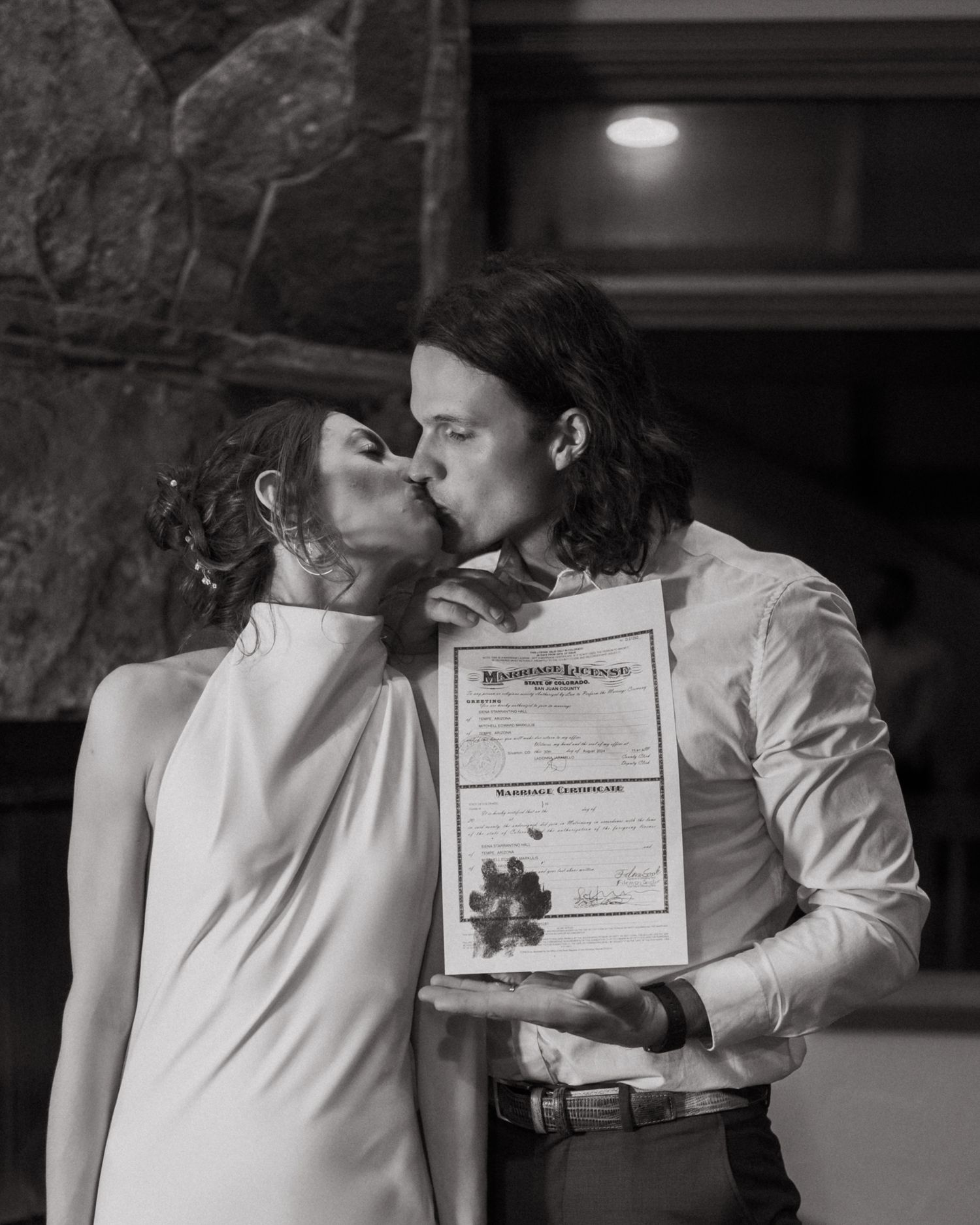 A couple shares a kiss while holding their marriage certificate in a rustic indoor setting.
