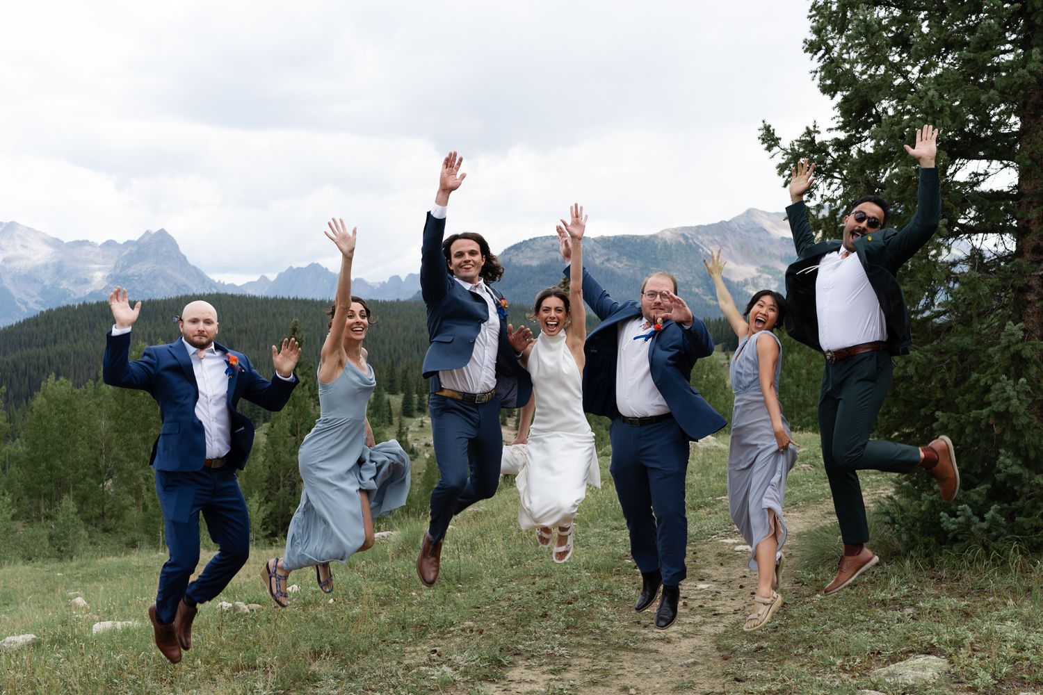 Wedding party jumps in celebration against mountain landscape backdrop.