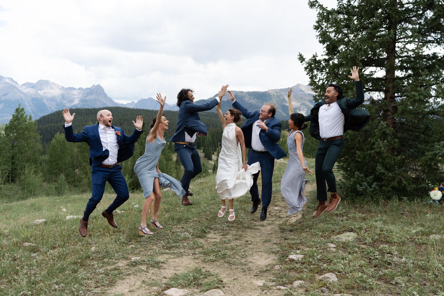Wedding party jumps joyfully against a backdrop of mountain peaks and evergreen trees.