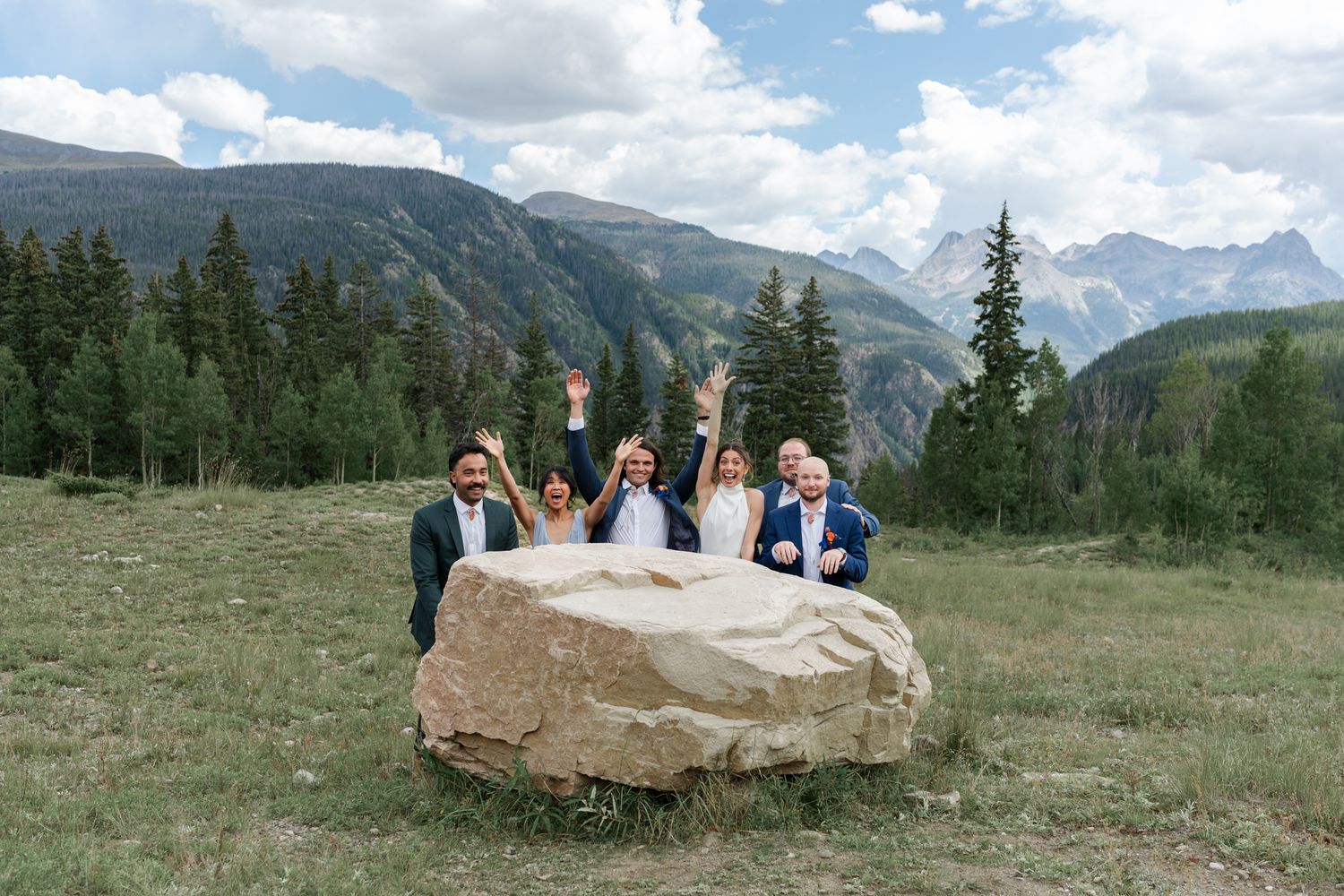 Wedding party gathers around large boulder with stunning mountain vista in background.