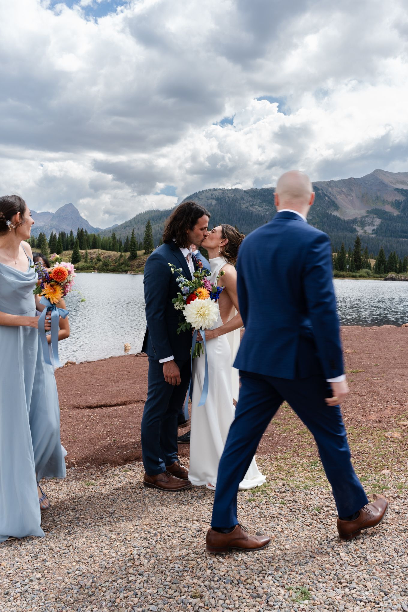 Wedding couple and guests gather by a mountain lake with dramatic cloudy skies.