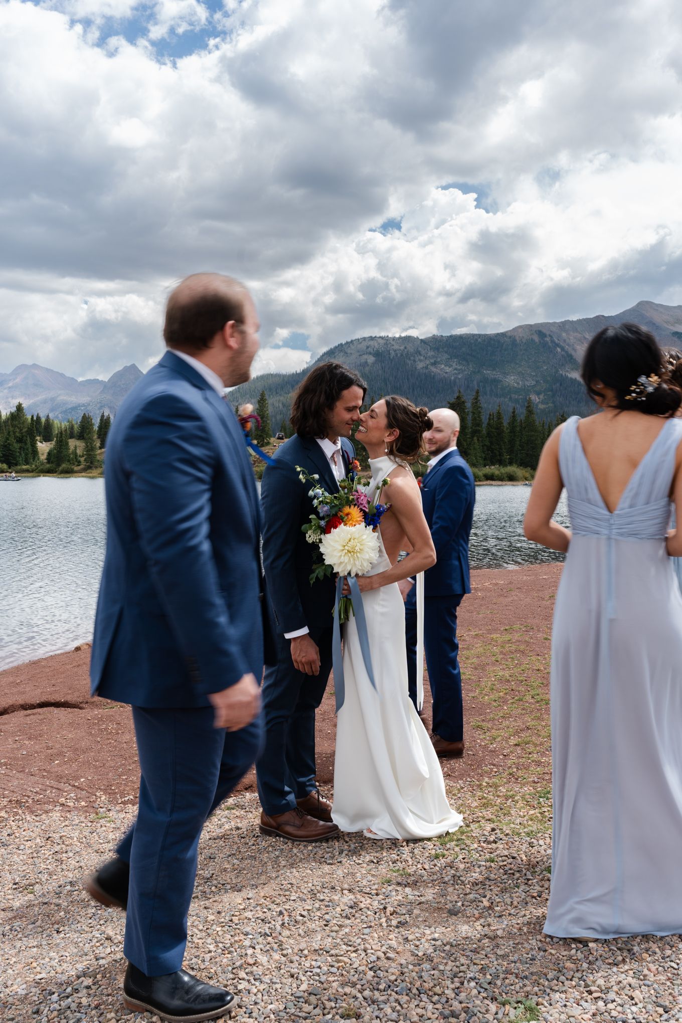 Intimate wedding moment captured by mountain lake with guests looking on.