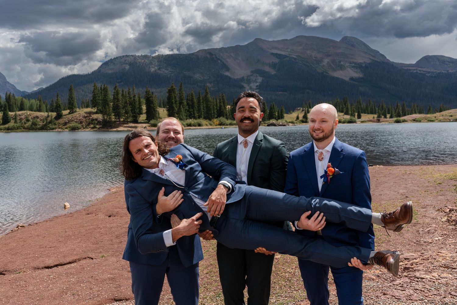 Playful wedding party lifts and carries someone in front of mountain lake.