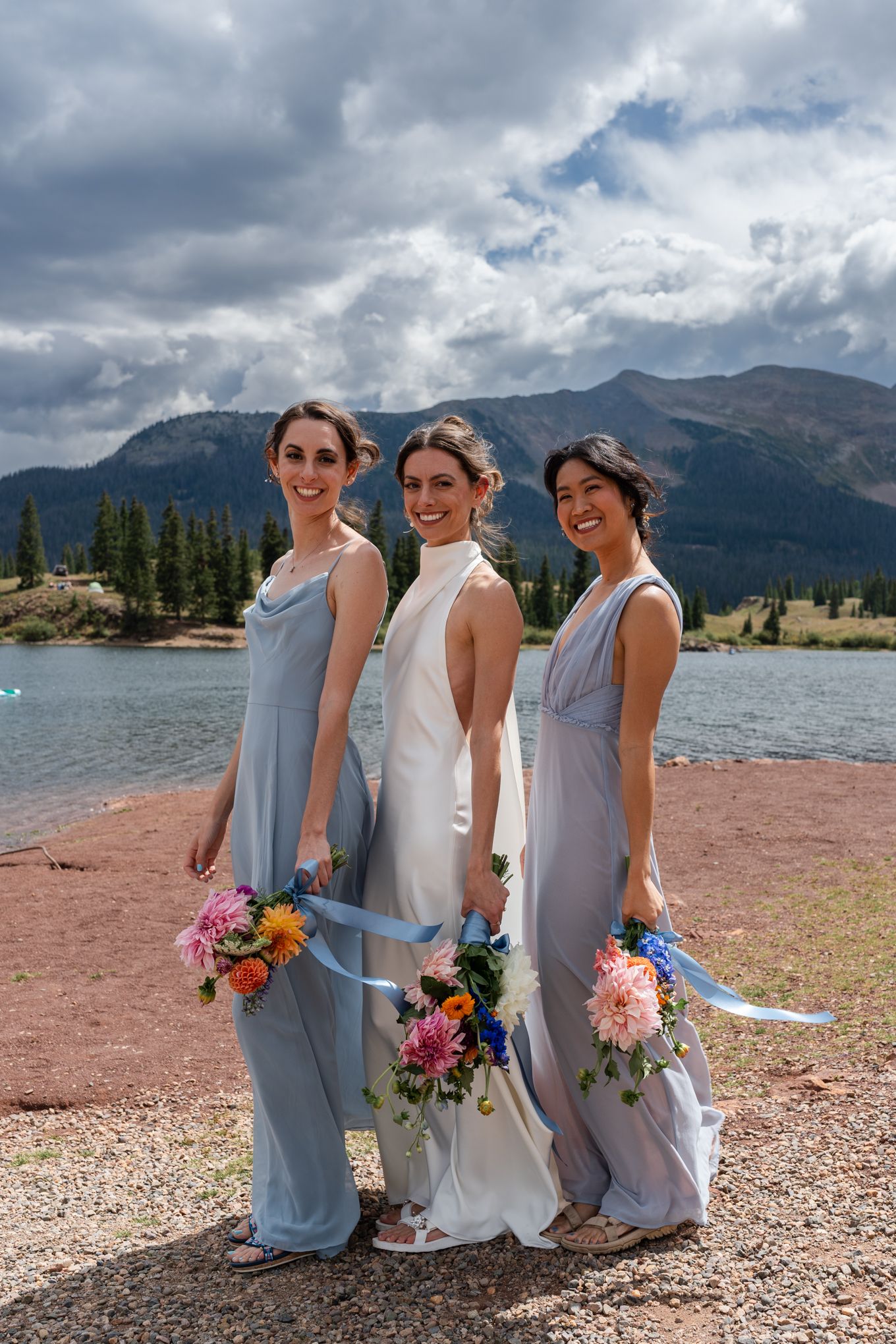 Three attendants in light blue dresses pose with bouquets by mountain lake.