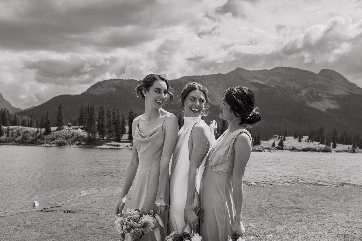 Three bridesmaids share a joyful moment against a dramatic mountain landscape in black and white.