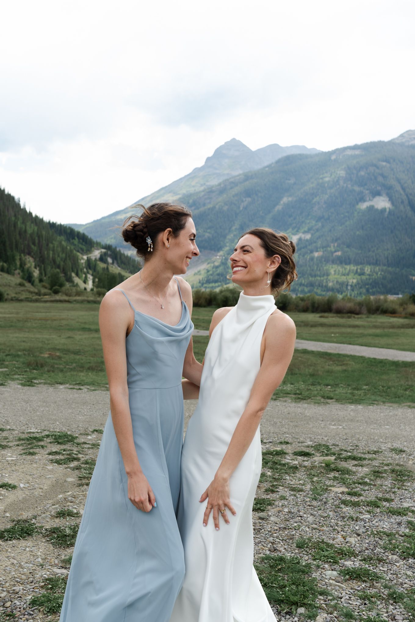 Two elegantly dressed figures share a candid laugh with mountains in the background.
