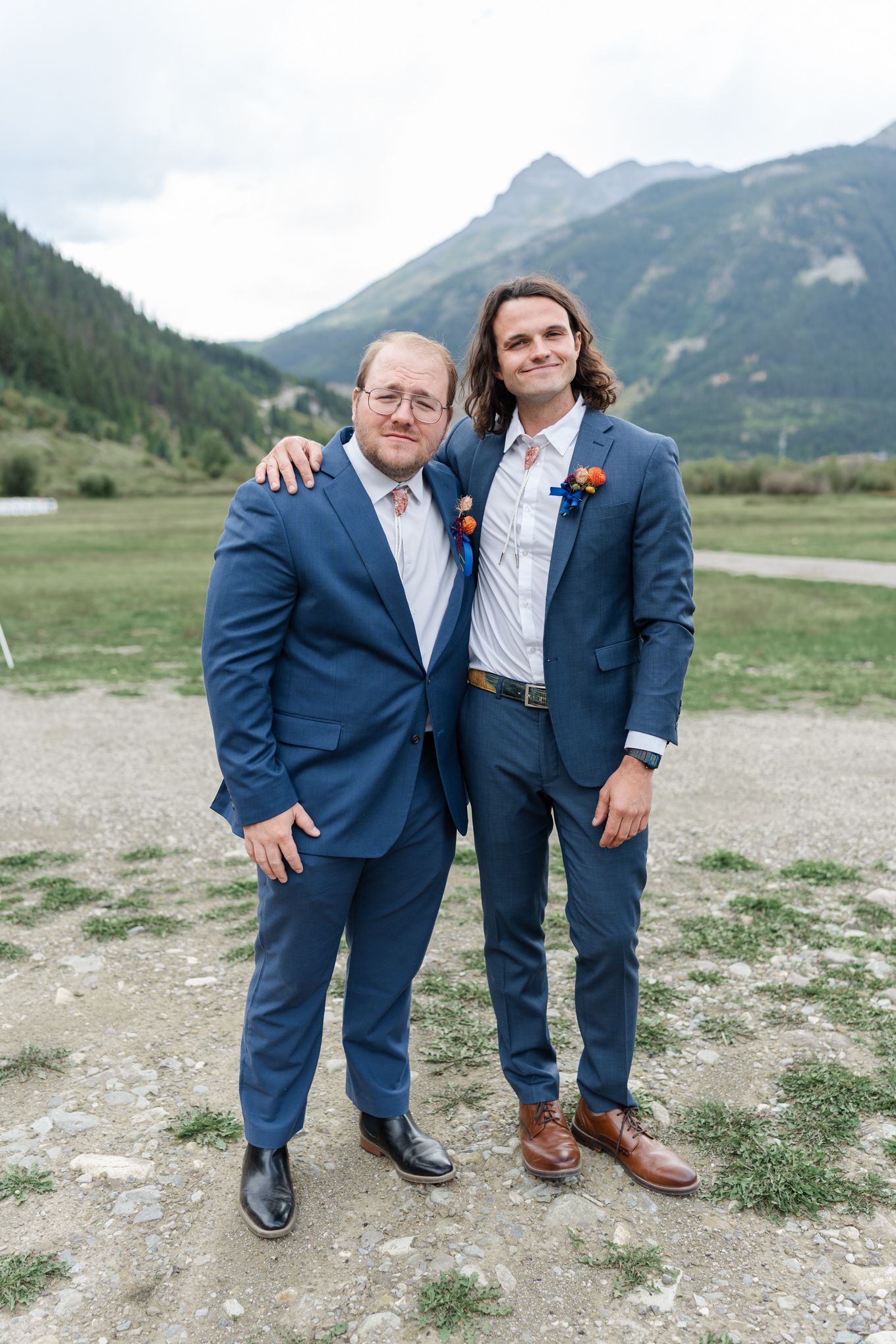 Two people in blue suits pose together against mountain backdrop.