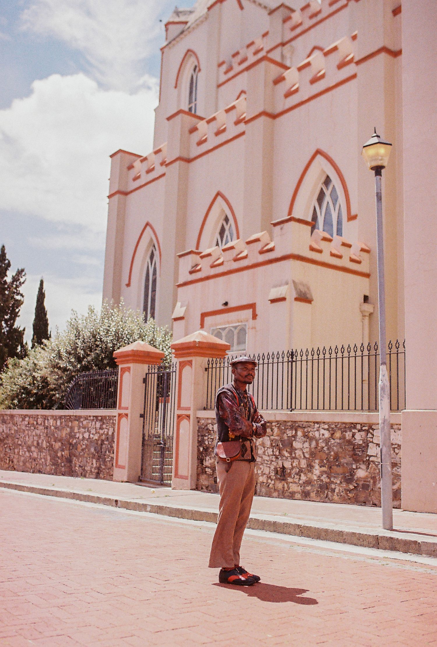 A vintage photograph shows a person standing on a sidewalk in front of a pink church with Gothic arches in Cape Town.