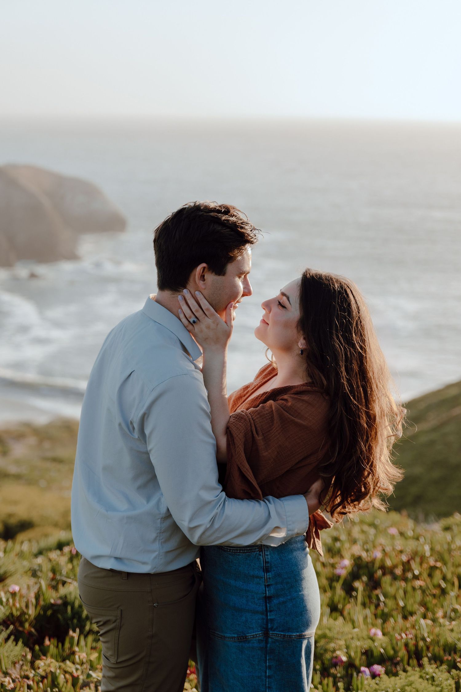 Romantic couple embracing on coastal cliffs at sunset with ocean waves and fog in background.