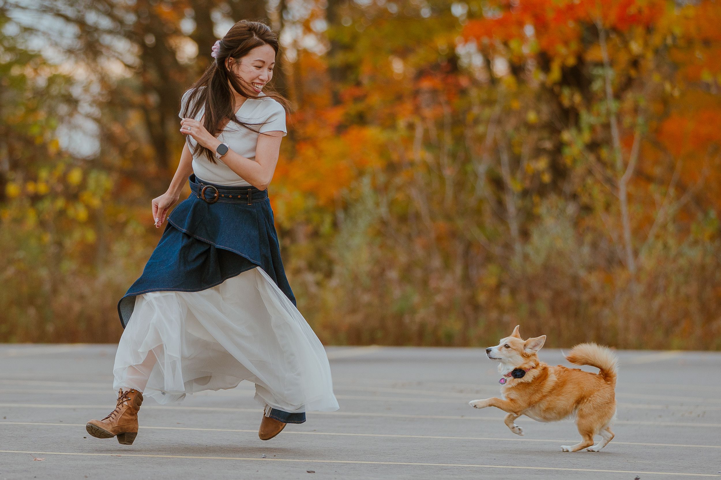 A person in layered clothing dances on a wooden deck with their Corgi dog against vibrant autumn foliage.
