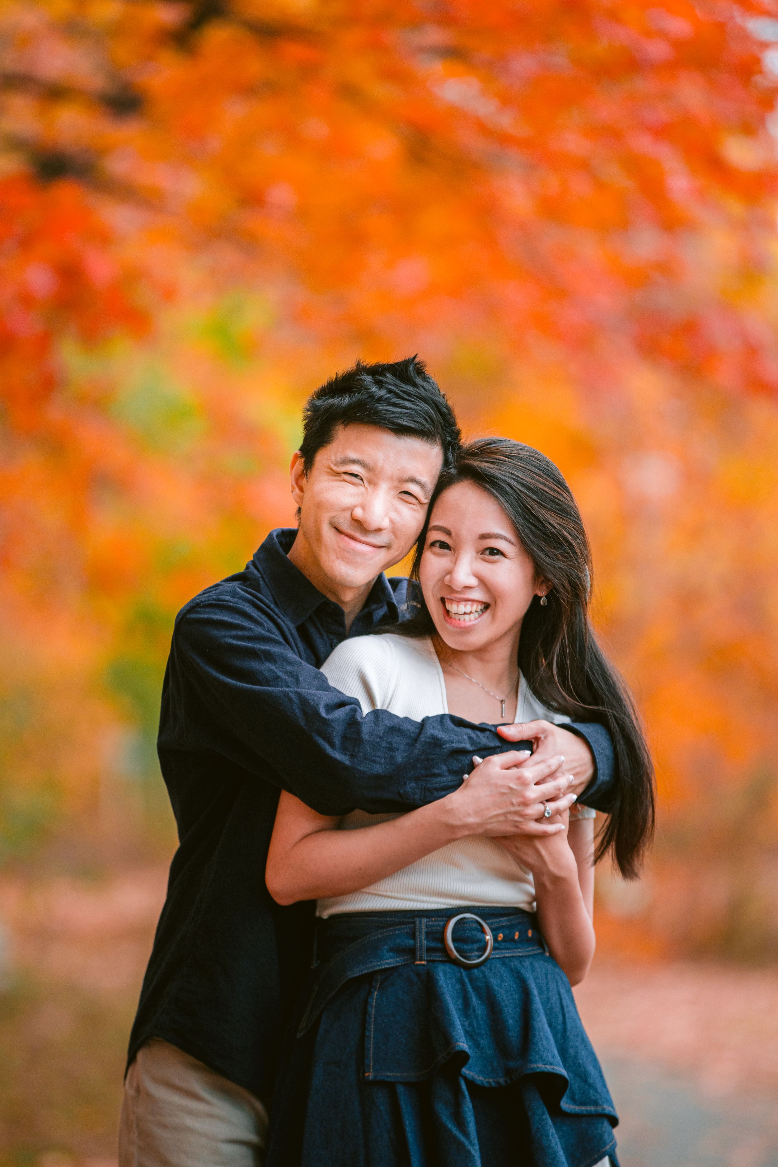 A couple embraces while laughing together against a backdrop of vibrant orange autumn foliage in a park setting.