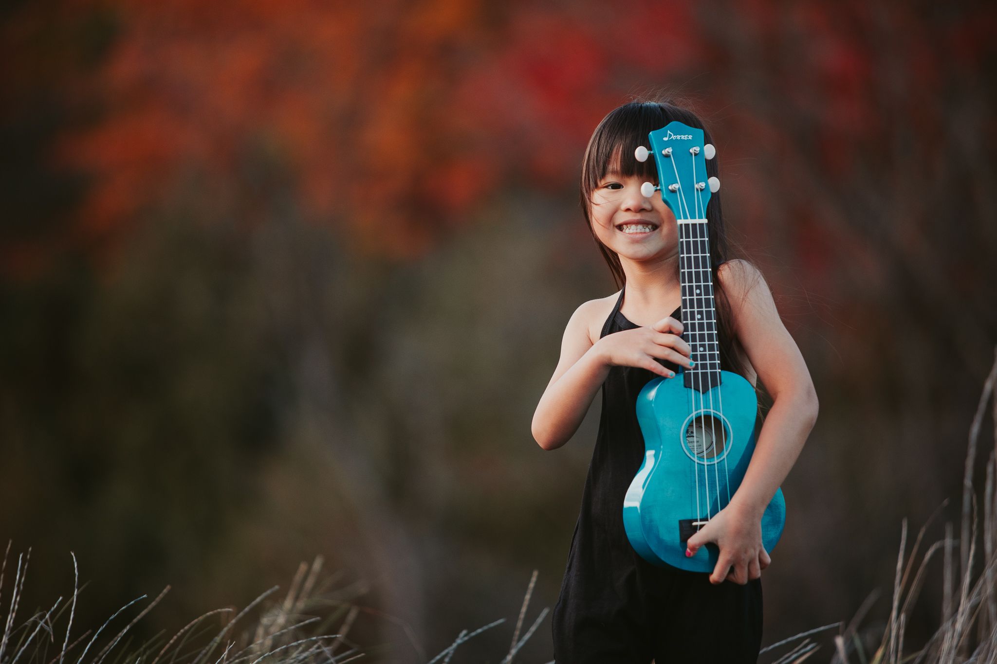 A happy child holds a turquoise ukulele while standing outdoors against a vibrant autumn background.