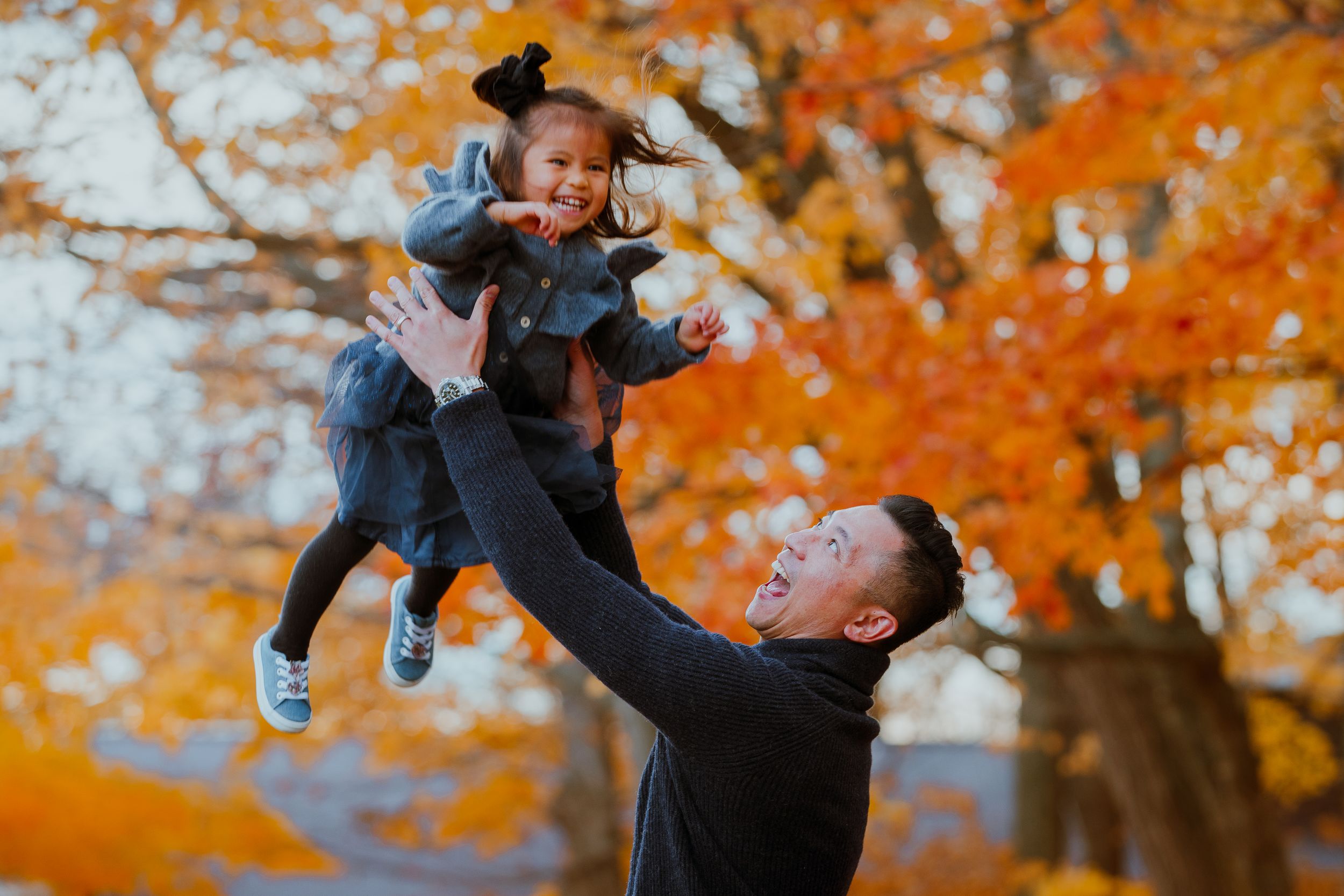 Playful family moment captured against vibrant orange autumn foliage as parent lifts child high in the air.