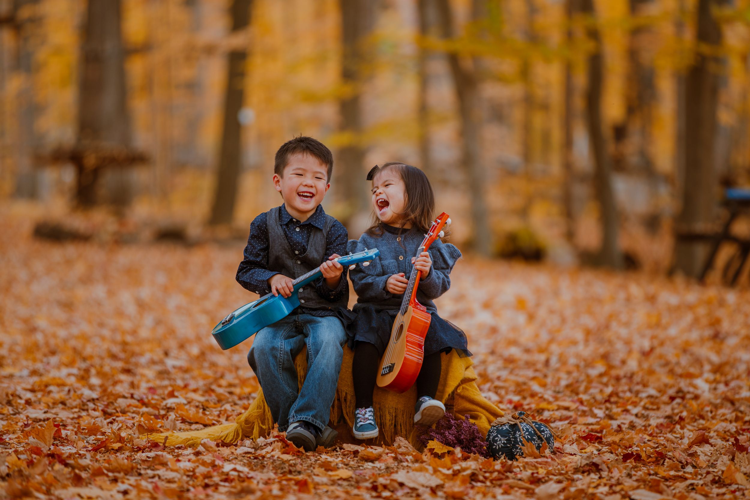 Two children sit on a log playing musical instruments surrounded by golden autumn leaves in a forest.