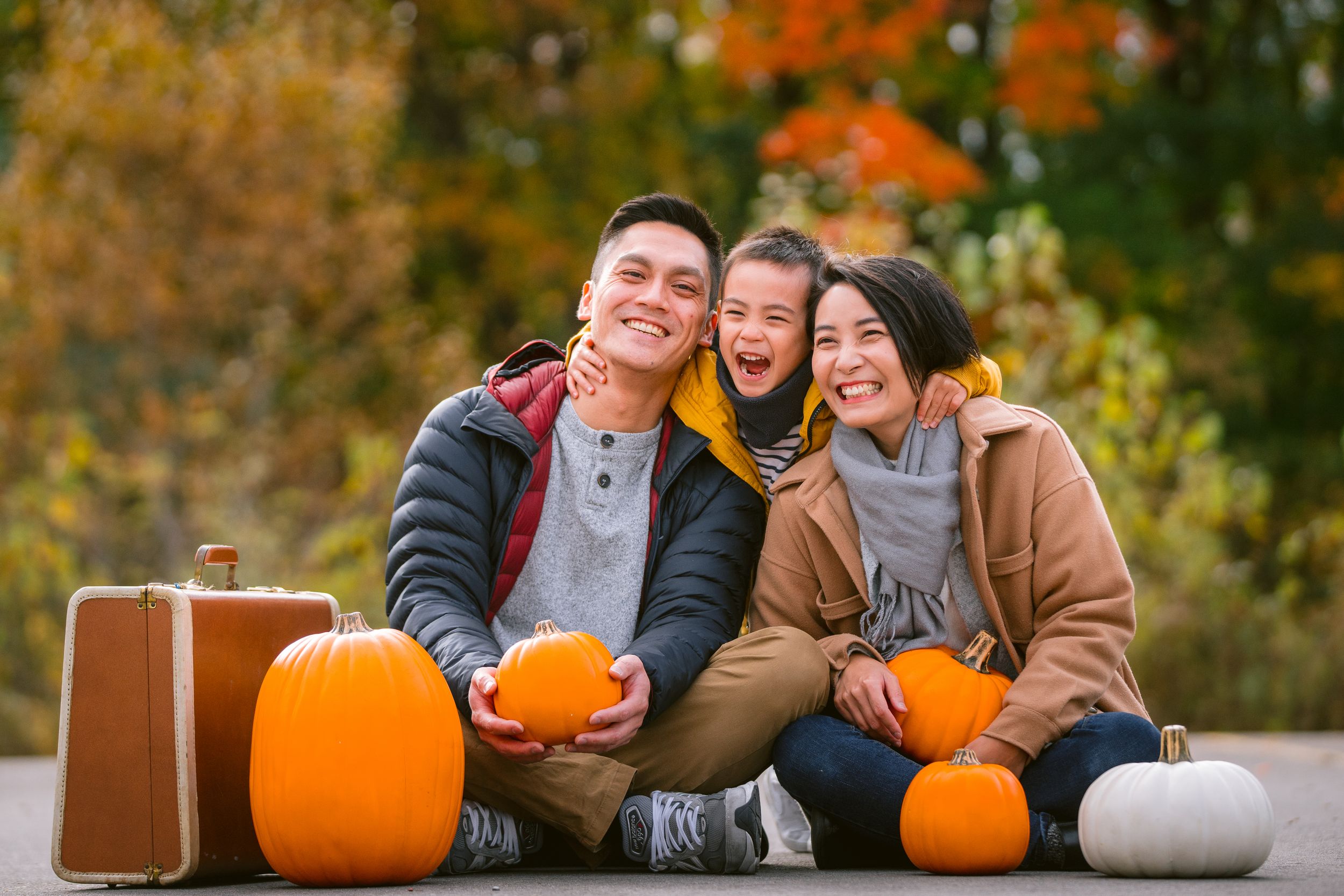 A happy family sits with pumpkins and vintage suitcase among fall foliage during an autumn outdoor photoshoot.