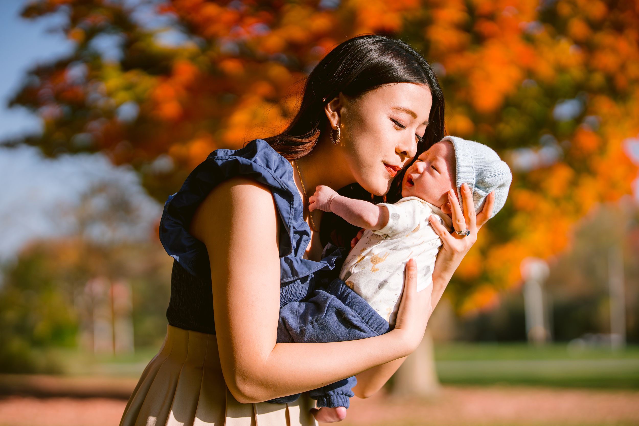 A mother tenderly embraces her baby outdoors against vibrant orange autumn foliage in the background.
