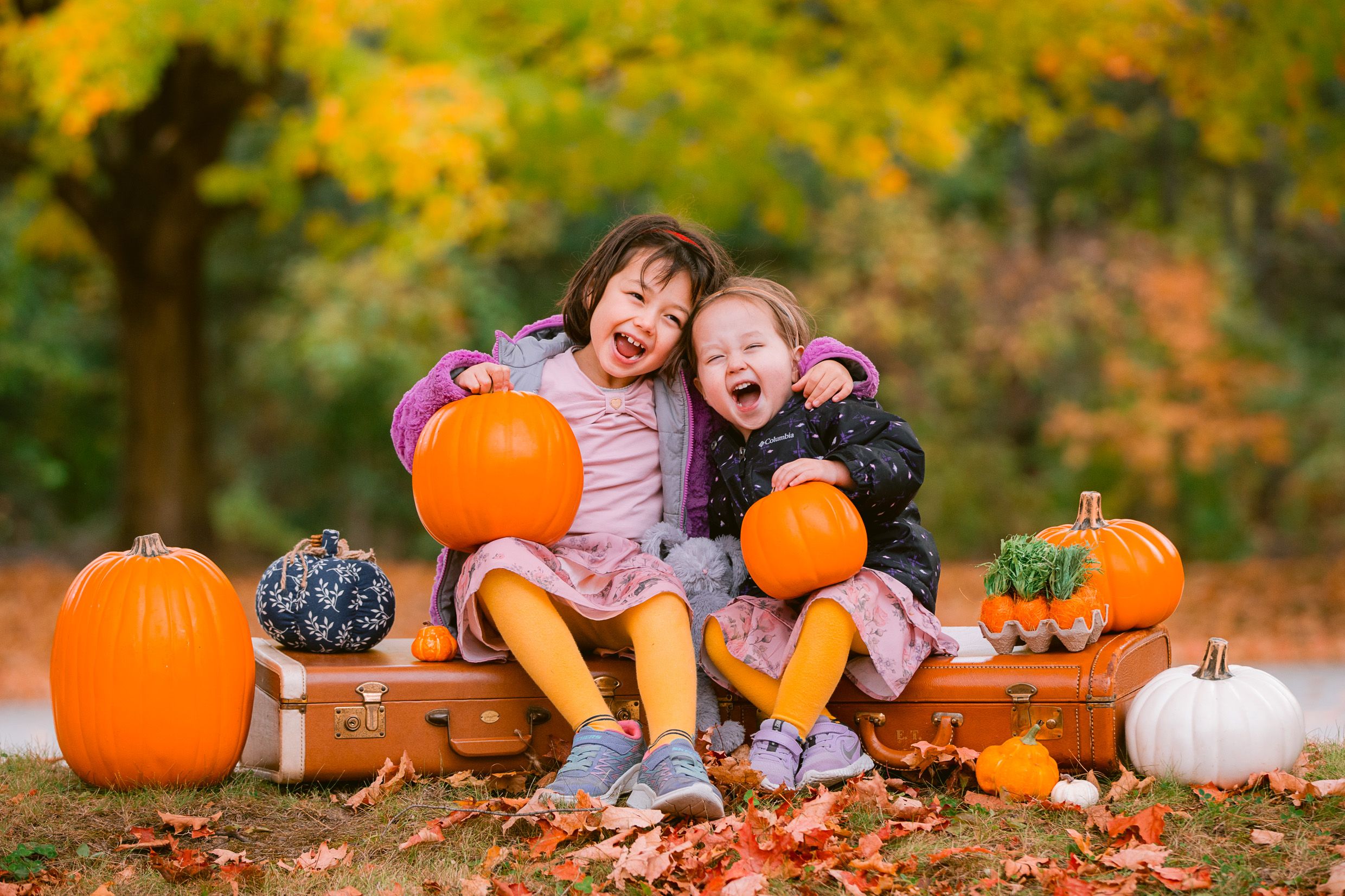 Children laugh while sitting with bright orange pumpkins and fallen autumn leaves in an outdoor fall setting.