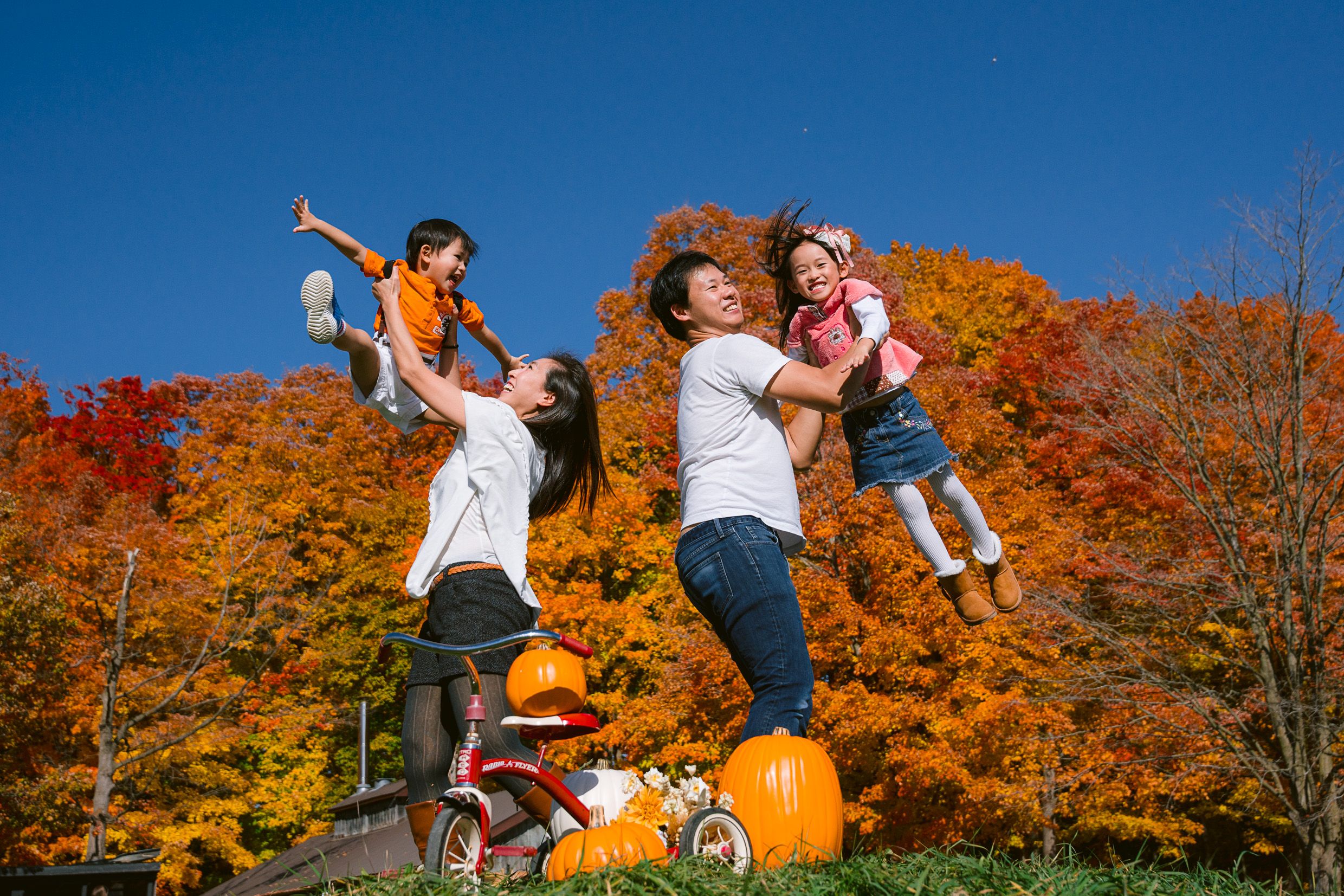 A joyful autumn scene with playful figures leaping in front of vibrant orange fall foliage and pumpkins on the ground.