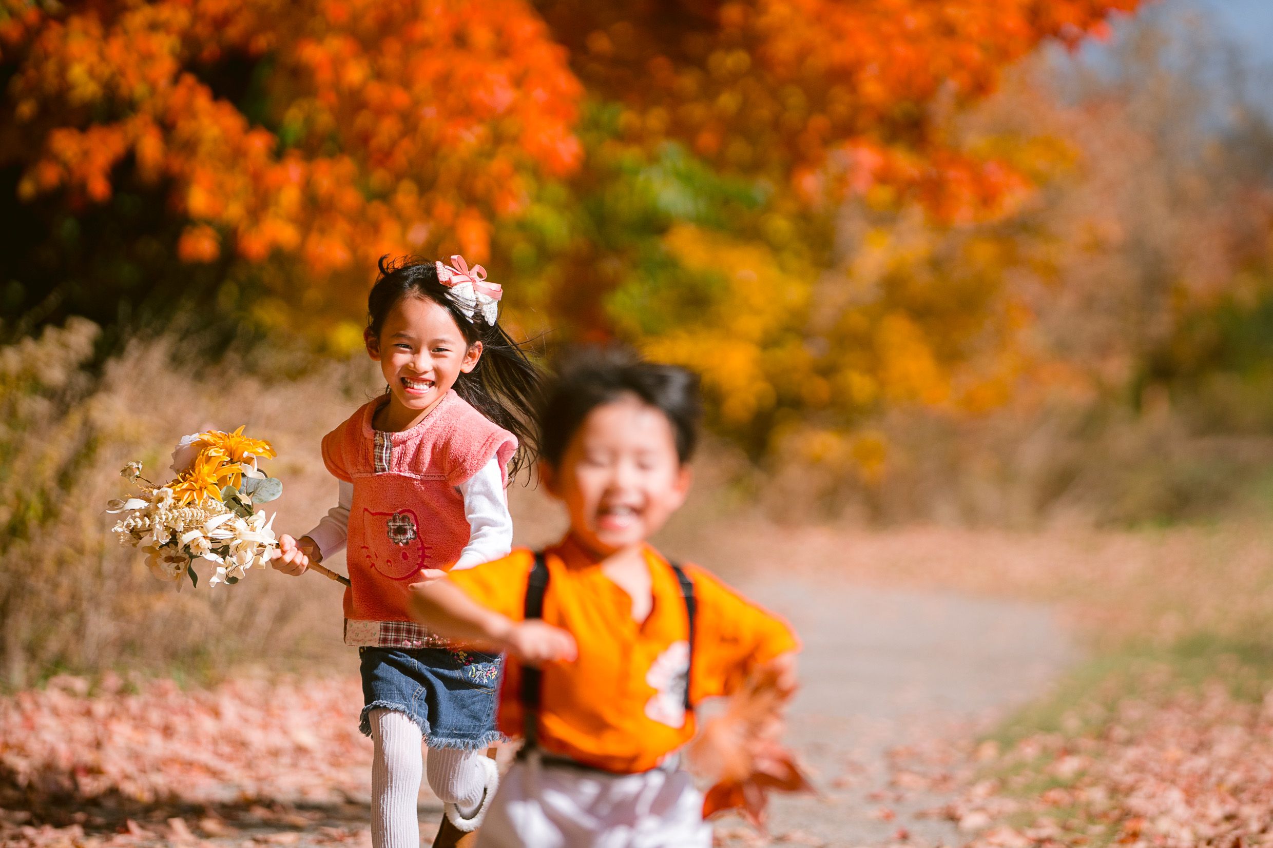 Young children running and playing with autumn leaves on a trail surrounded by vibrant orange fall foliage.