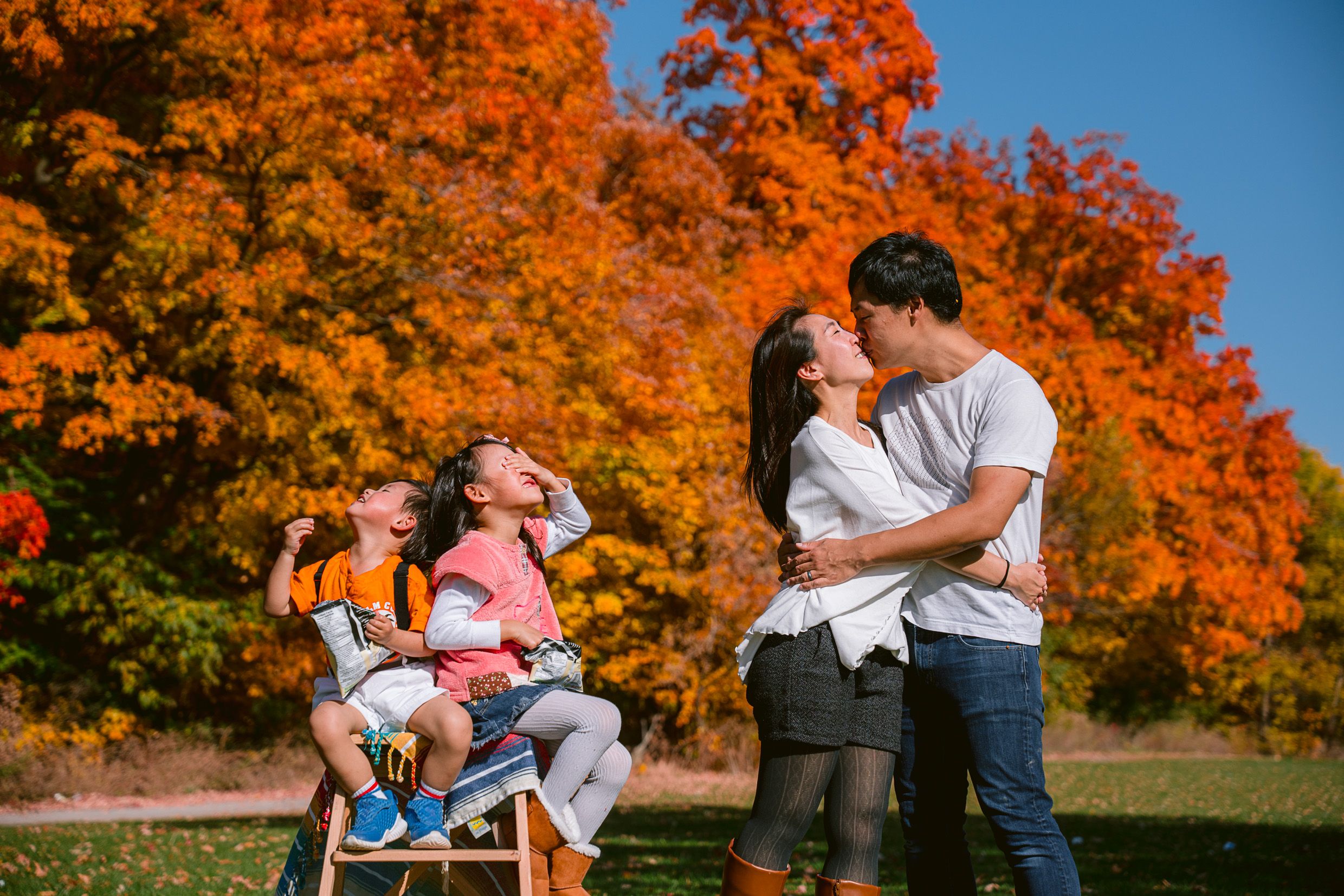 Couple embracing in a park with vibrant orange autumn foliage while children play in background on sunny day.