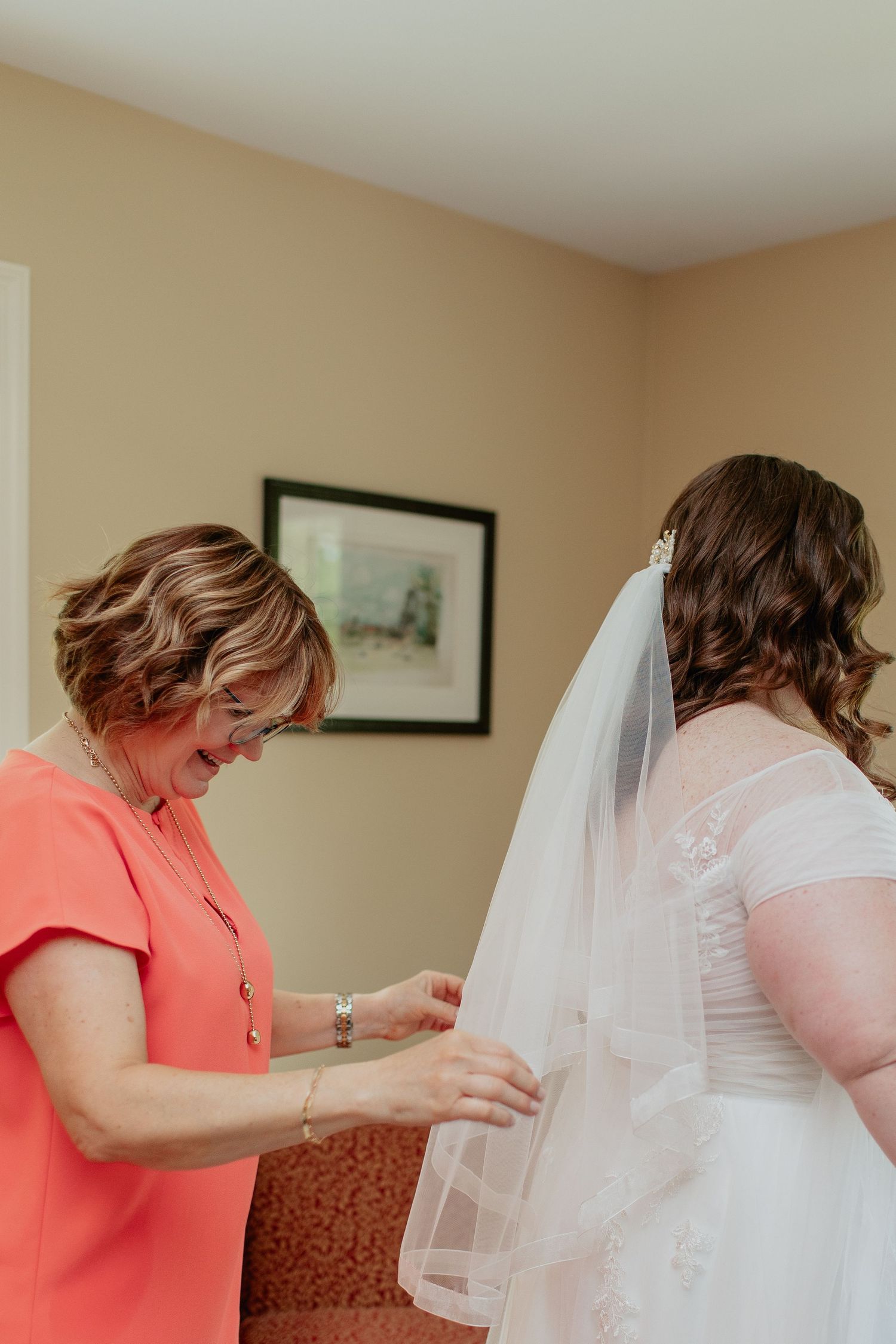 Someone in a bright coral dress helps adjust a wedding veil on a bride wearing a white lace dress.