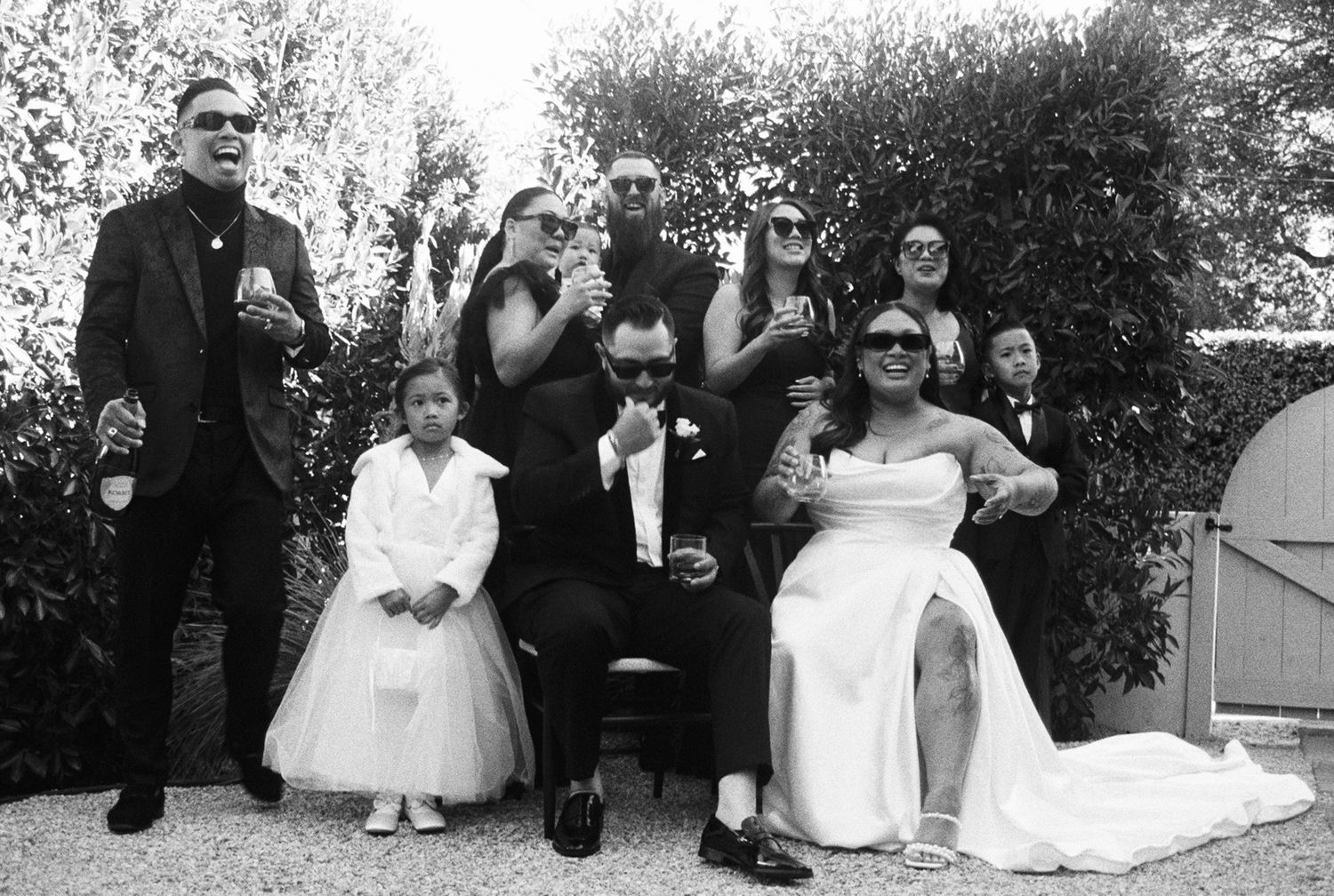 A black and white photograph showing a wedding party group posing together outdoors by bushes.