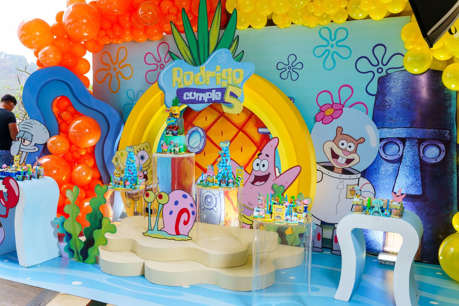 Colorful children's birthday party table decoration with cartoon character theme featuring bright rainbow backdrop and balloons.
