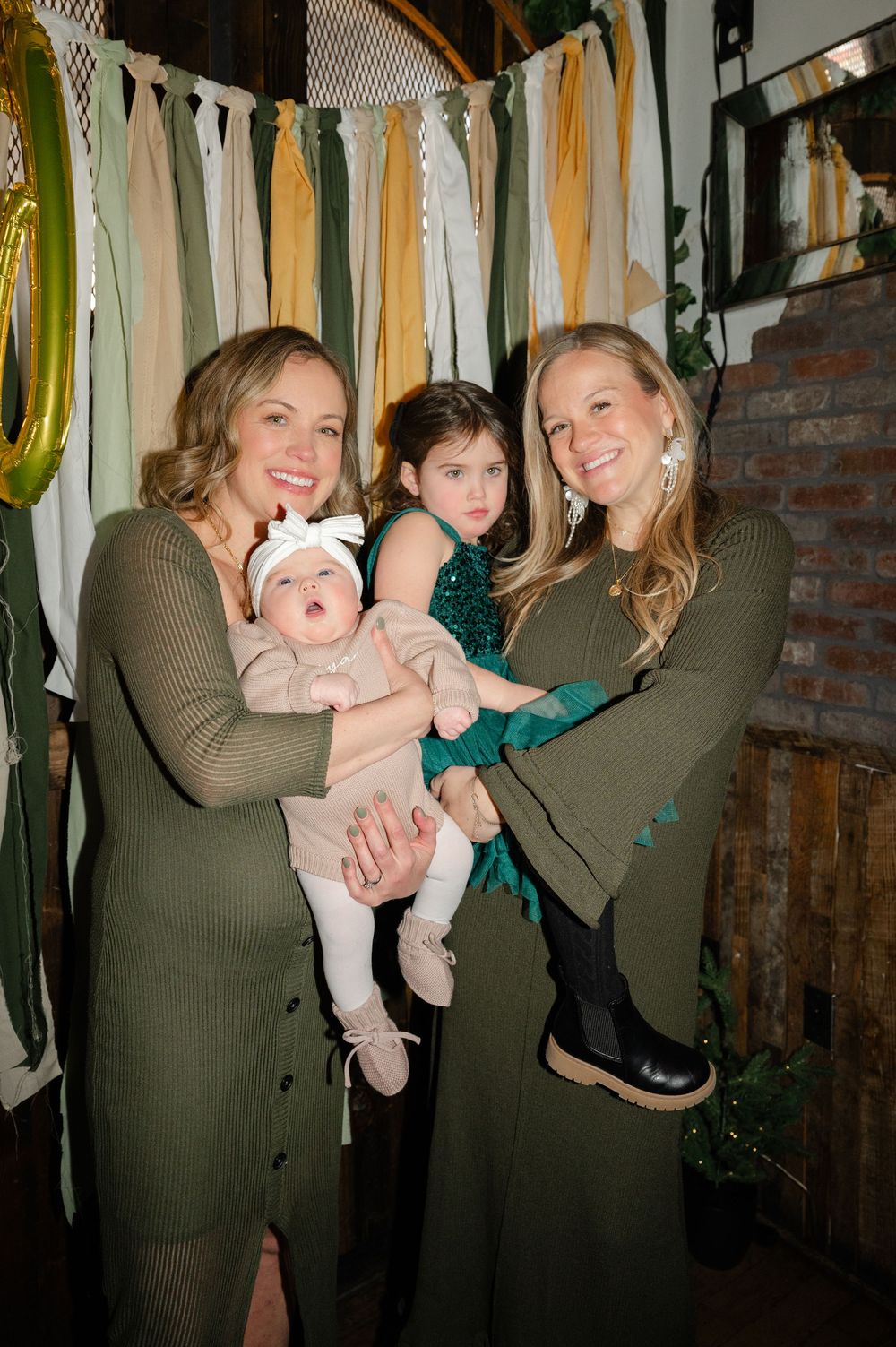 Two women in olive sweaters pose with two children against a backdrop of hanging fabric strips in neutral tones.