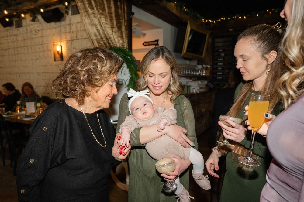 A social gathering at a dimly lit venue where people are chatting and admiring a baby dressed in pink.