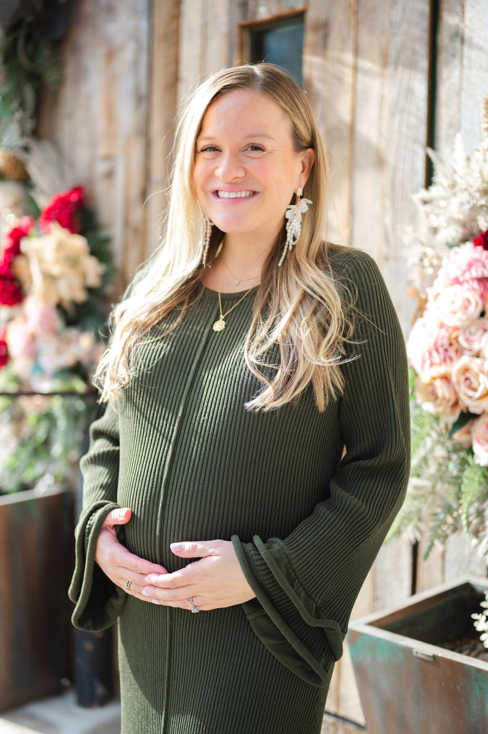 Expectant mother in olive green sweater dress poses in front of floral decor for maternity photo.