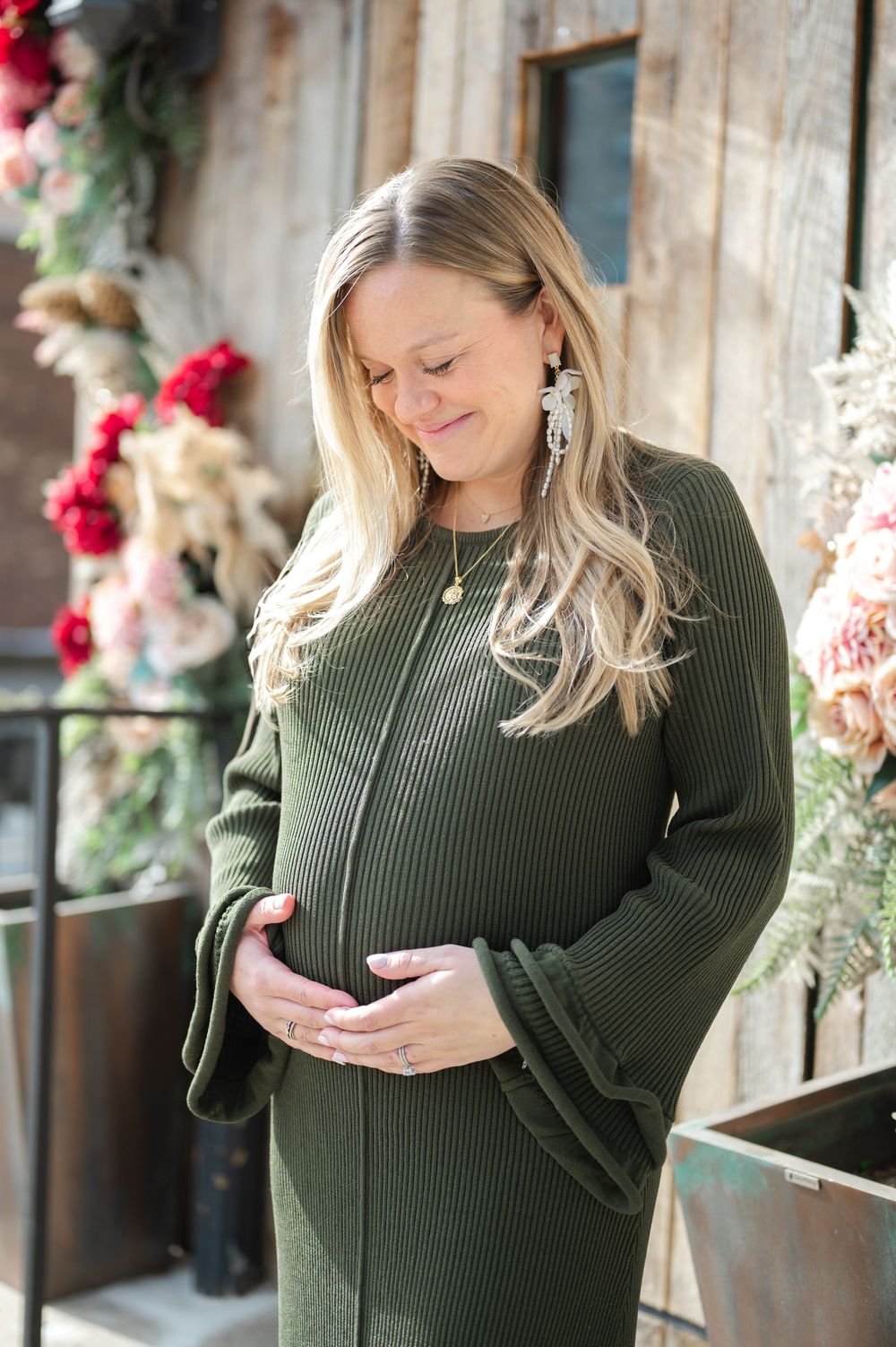 Expectant mother in olive green sweater dress stands amid floral decorations with a gentle smile.