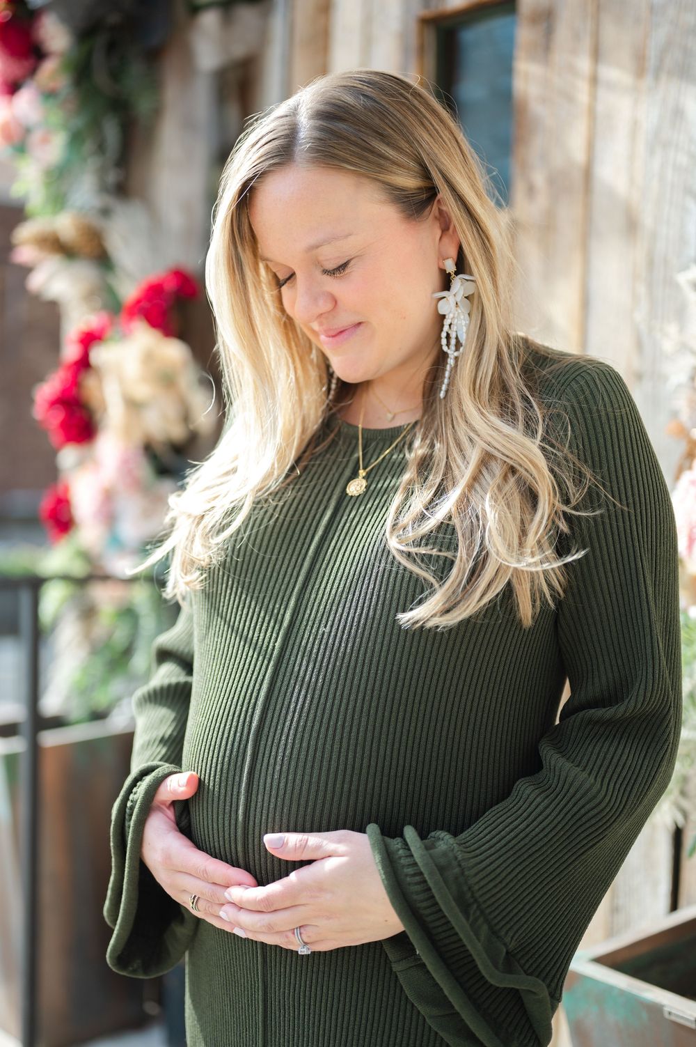 An expecting woman in a green ribbed sweater with bell sleeves gently touches her baby bump outdoors.