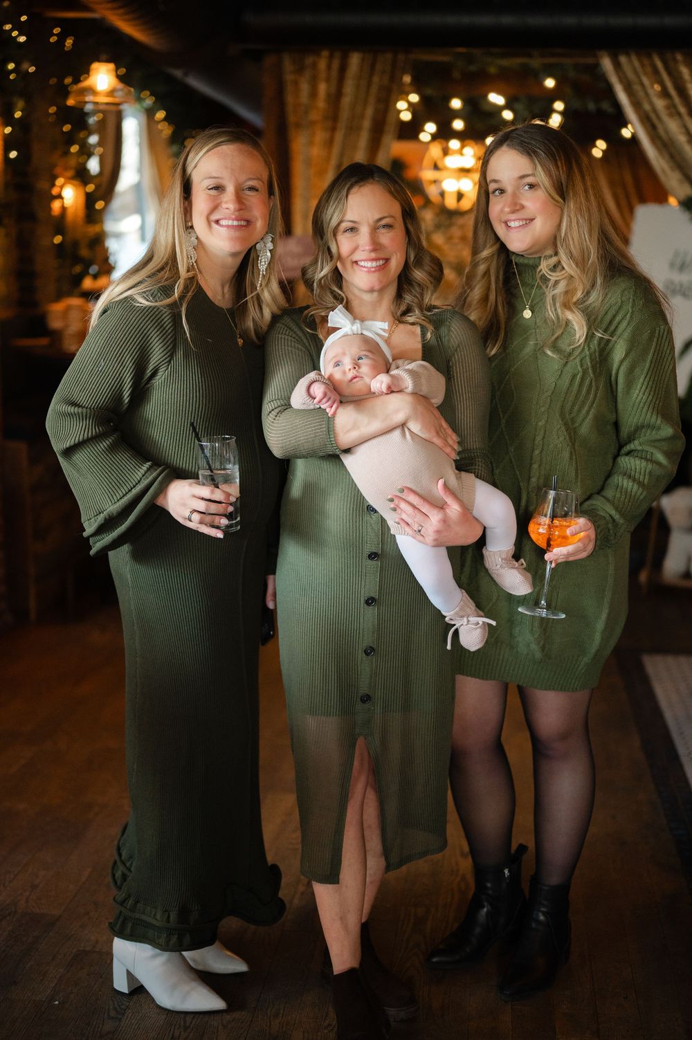 Three ladies in coordinating olive green dresses pose with a baby at an elegant indoor event.