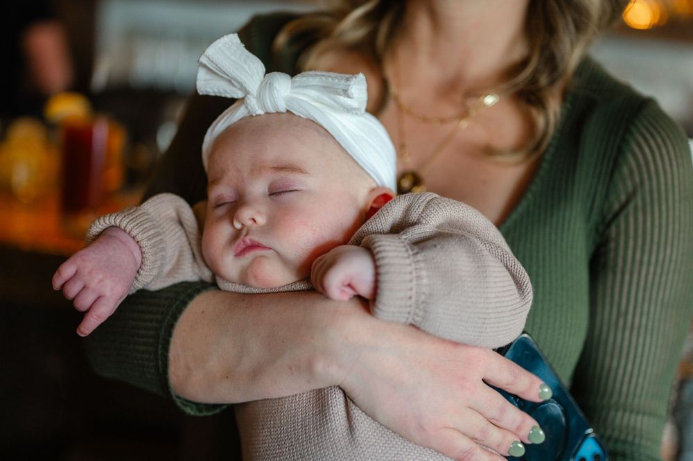 A sleeping baby wearing a white bow and beige sweater rests peacefully in arms against a green sweater.