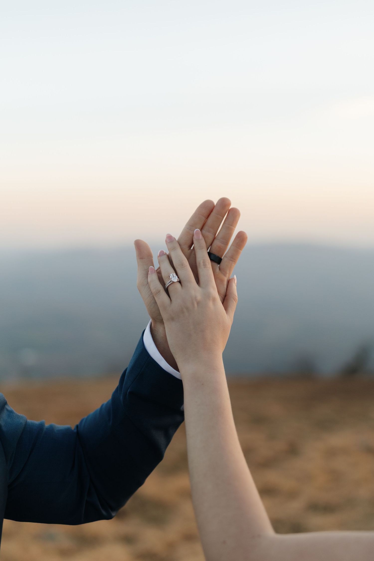 Two hands with wedding rings touching against a blurry mountain sunset background.