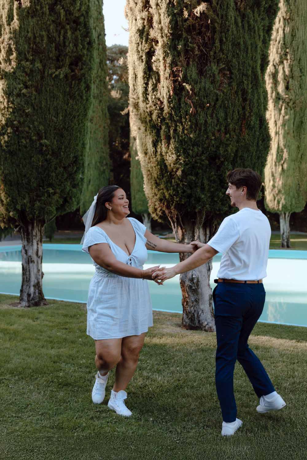 Two people in white outfits dancing together on grass near a pool and cypress trees at dusk.