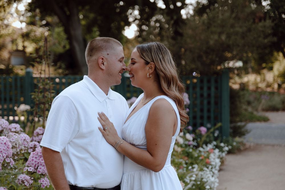 A romantic couple in white outfits share an intimate moment on a garden path surrounded by pink flowers and greenery.