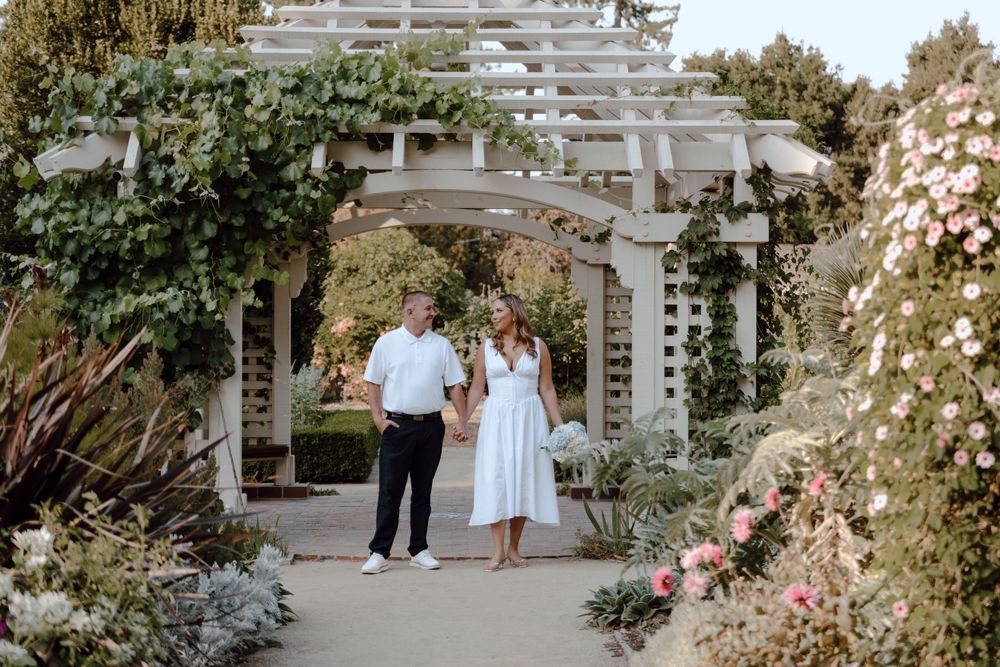 A couple stands beneath a white garden pergola surrounded by climbing roses and lush landscaping in romantic garden setting.